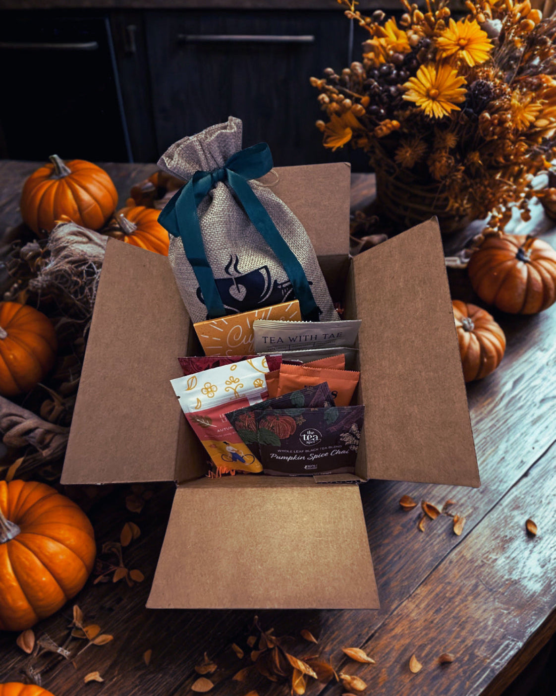 An open fall collection gift box filled with assorted packaged tea and gift items, styled on a wooden table with pumpkins, dried flowers, and autumn leaves.